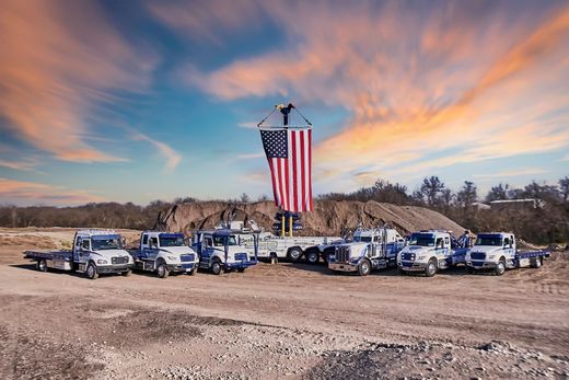 Line of white utility trucks beneath a giant American flag at sunset on a dusty lot.