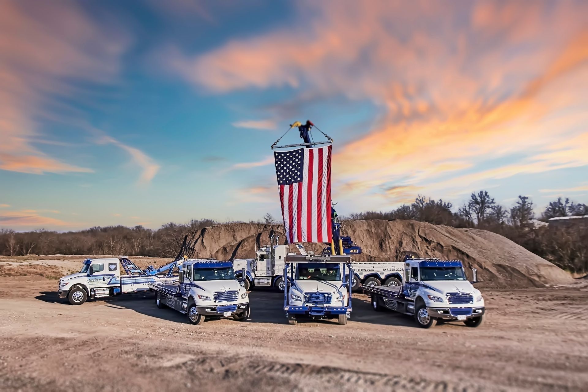 Line of utility trucks beneath a large American flag at a worksite at sunset