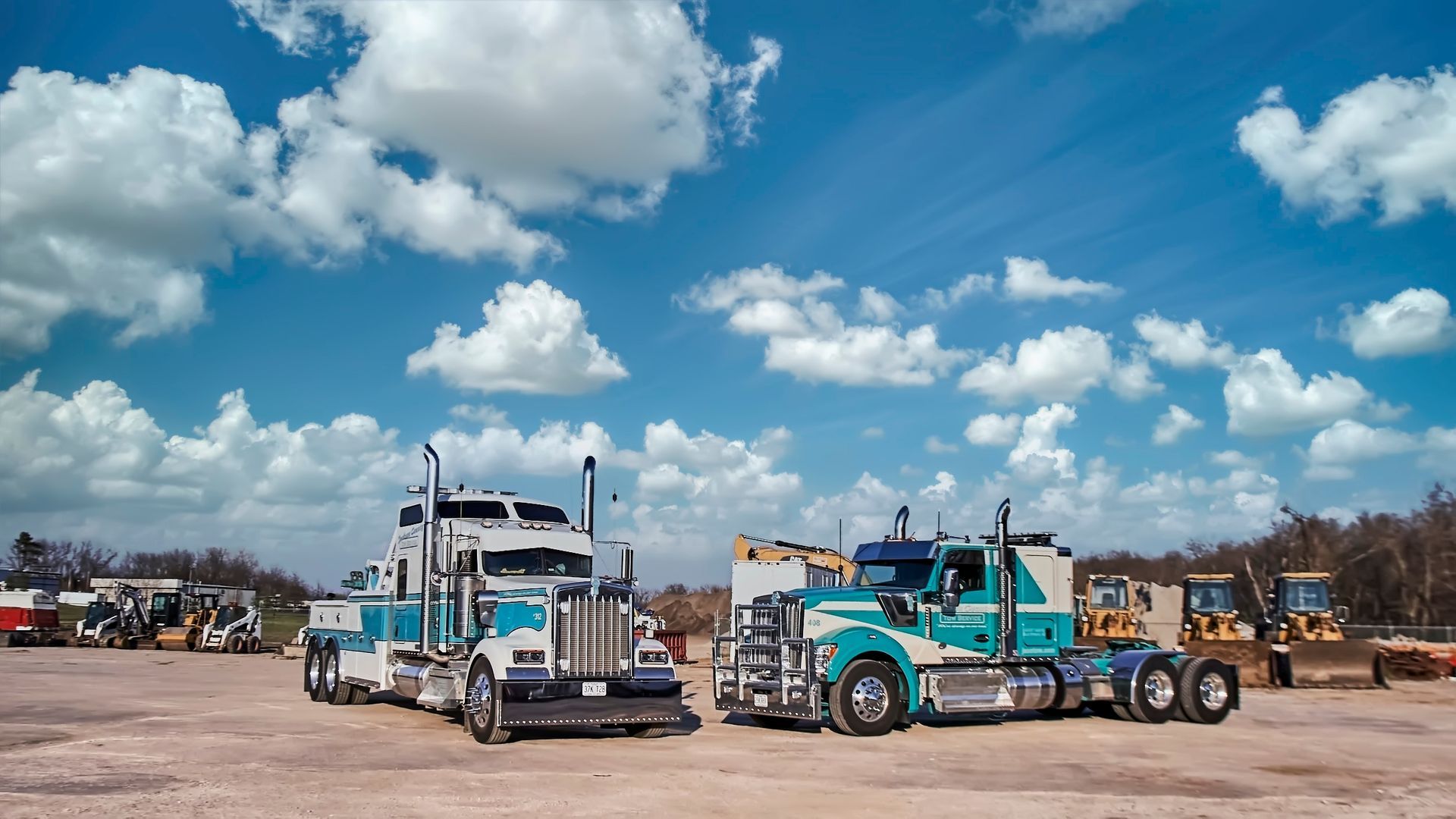 Two turquoise semi trucks parked on a dusty lot under a bright blue sky with clouds