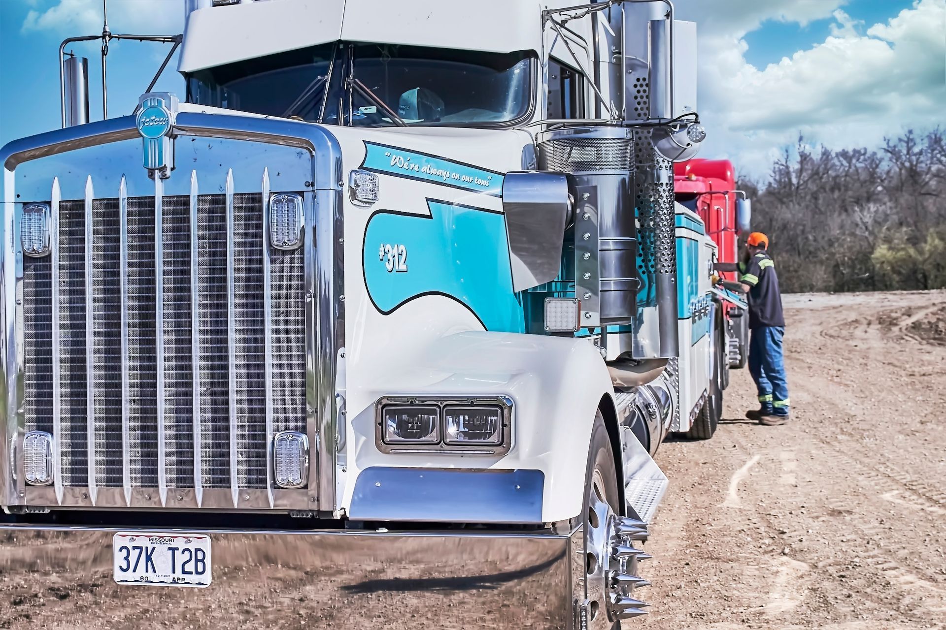 Blue and white semi truck parked on a dirt road, with a man standing beside it under a cloudy sky