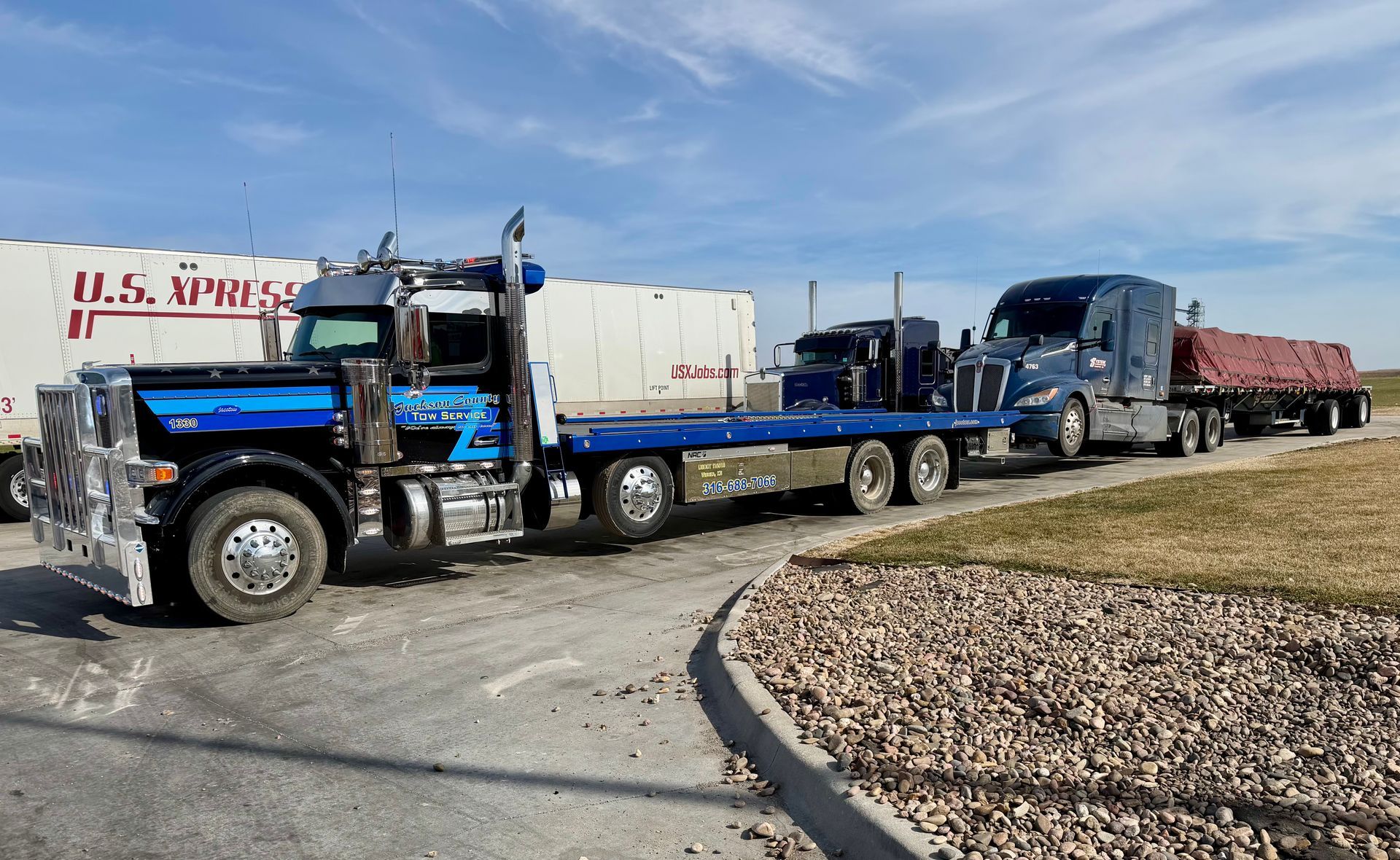 Blue semi-truck hauling a long flatbed trailer with a second truck, parked on a paved lot.
