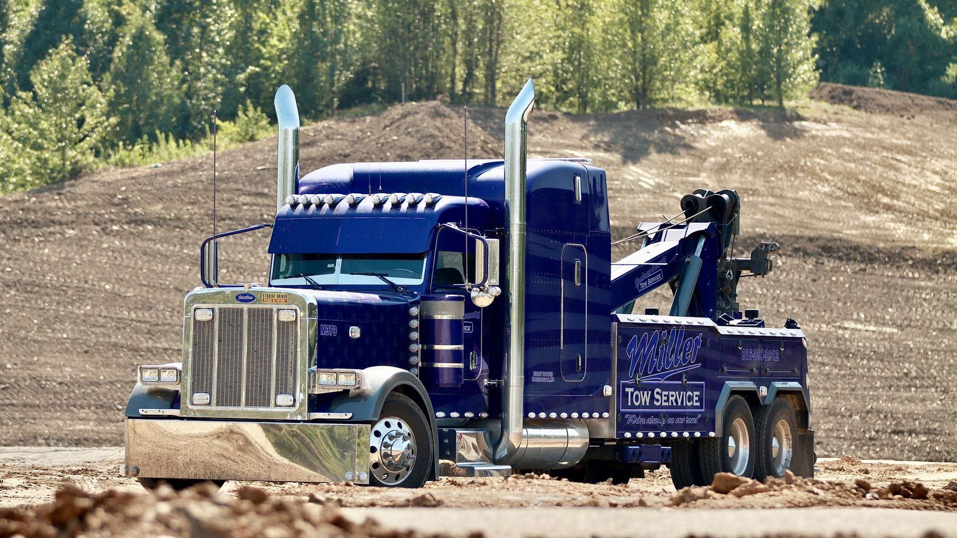Blue semi-truck hauling a black boom crane on a dirt road with trees and hills in the background