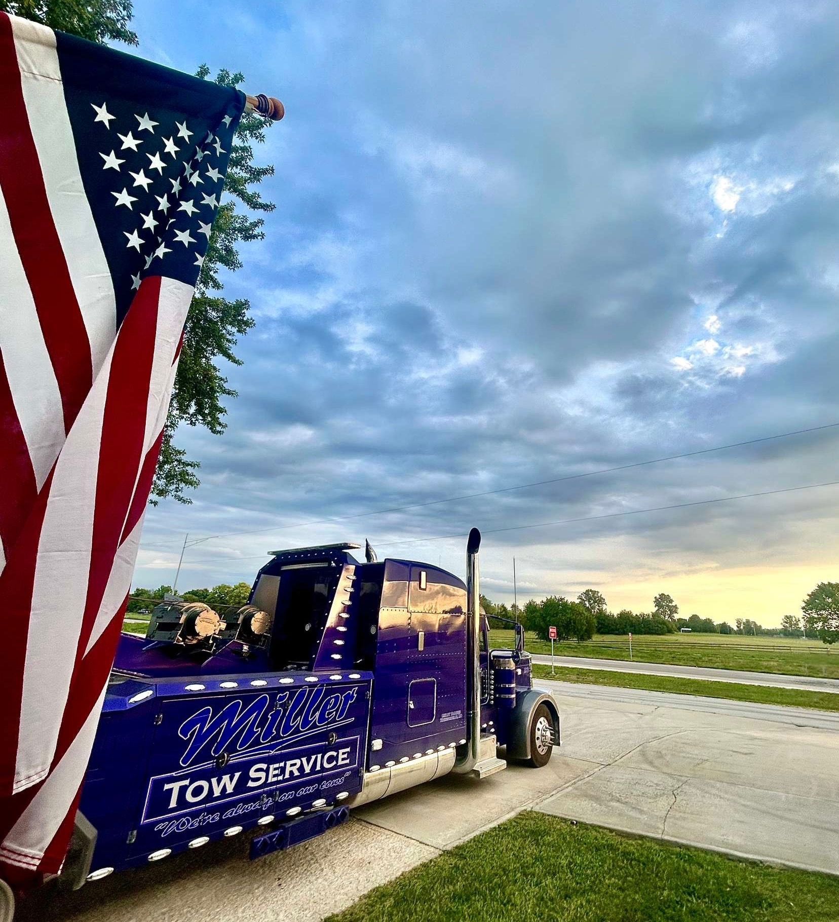 Blue tow truck with U.S. flag parked on driveway under cloudy sky at sunset