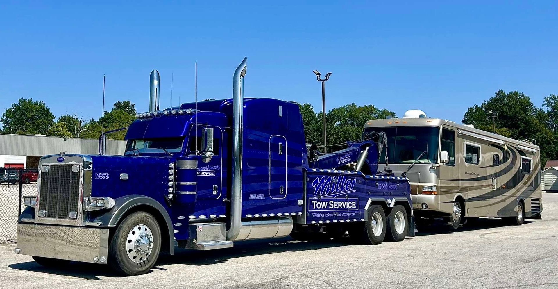 Blue semi-truck hauling a silver RV on a gravel lot under a clear sky