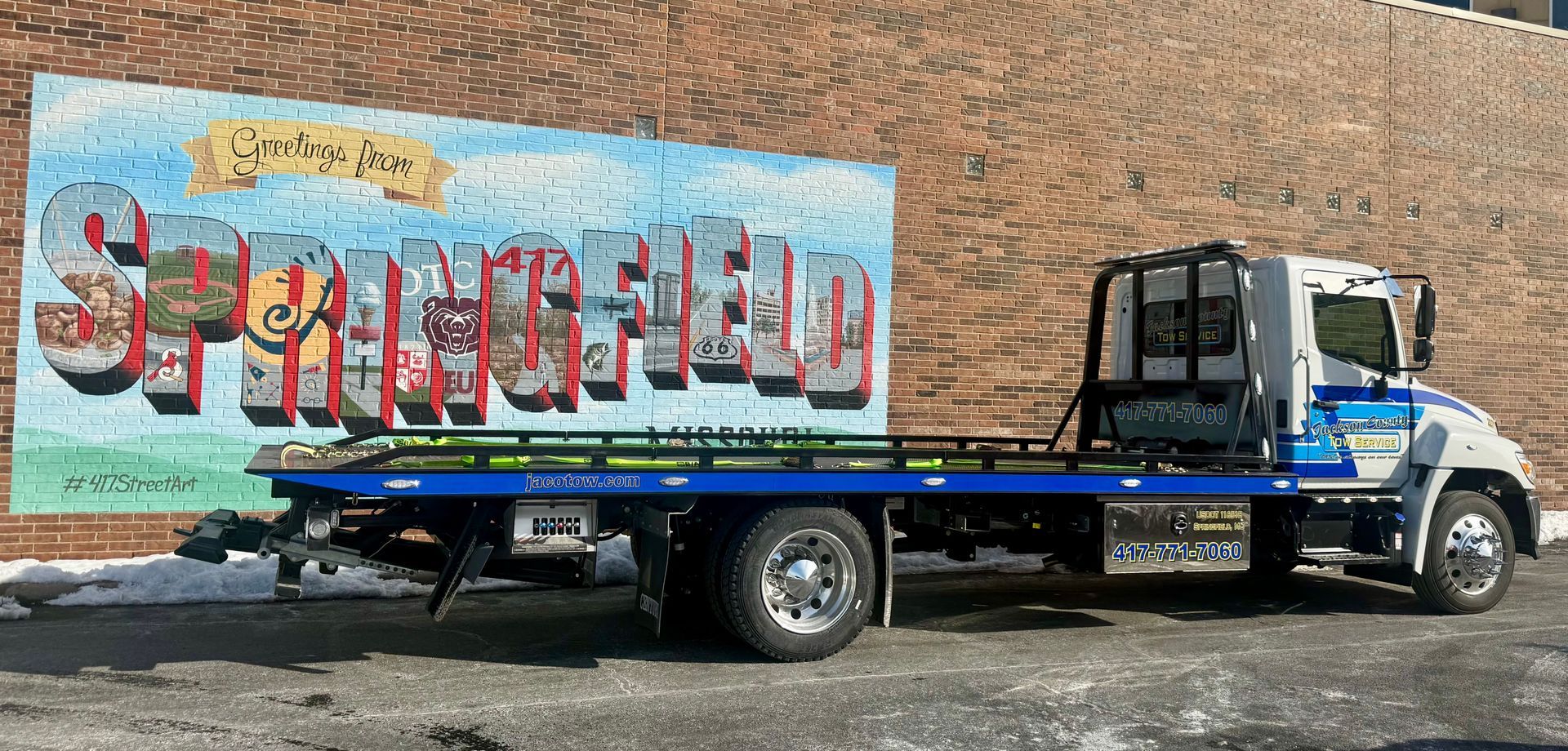 Flatbed tow truck with a large graffiti mural on the side, parked beside a brick wall.