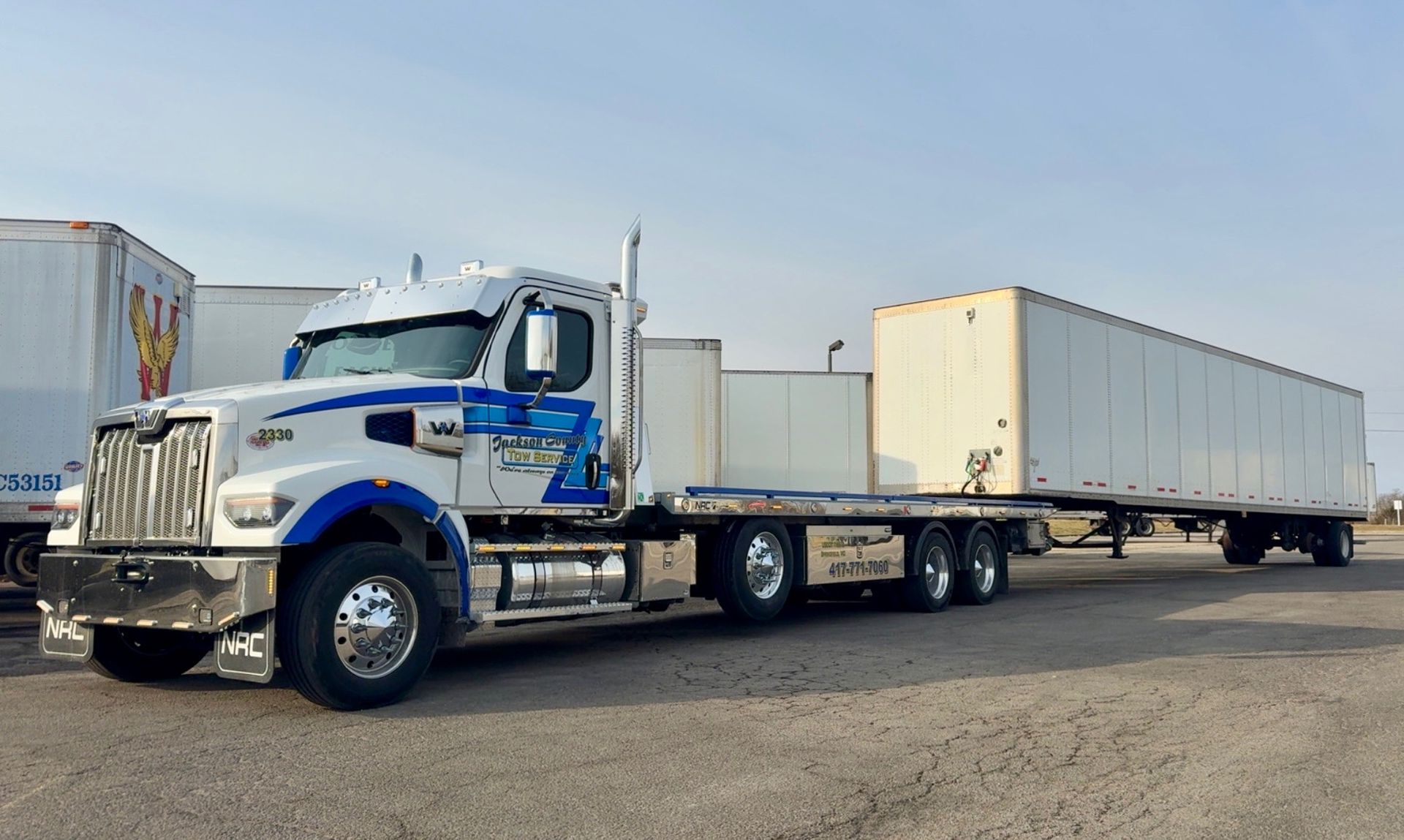 Blue semi truck hauling two white trailers in a yard under a clear sky