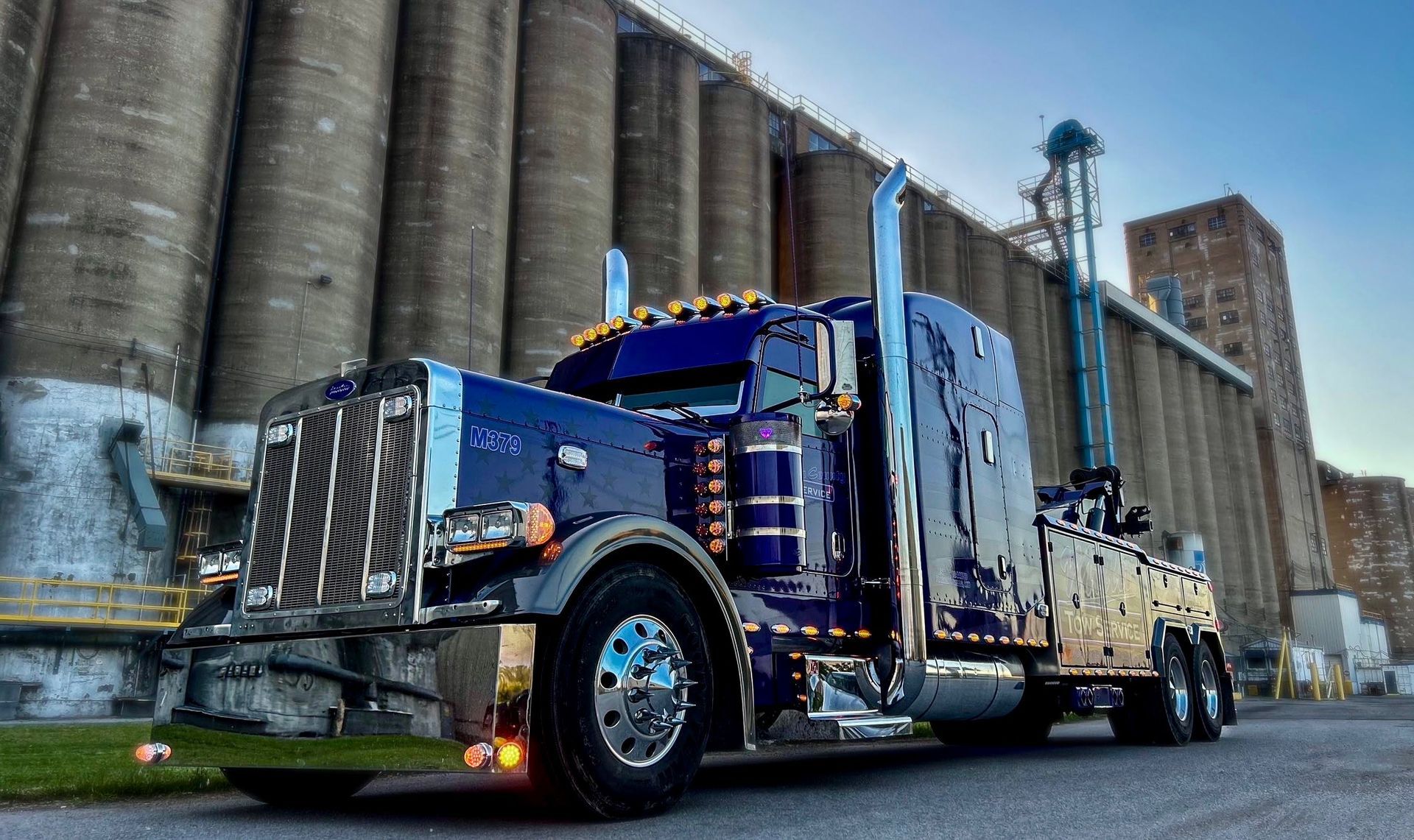 Blue semi truck parked beside tall industrial silos and a brick building.