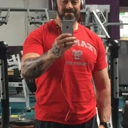 A muscular person wearing a red t-shirt and headphones takes a mirror selfie in a gym setting.