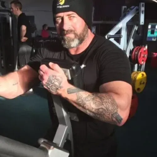 A bearded person in a black beanie and t-shirt with tattooed arms exercises on a machine in a gym.
