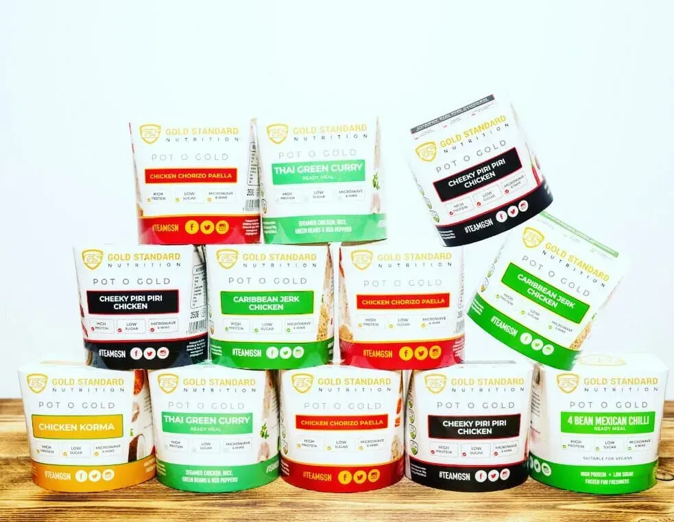 A pyramid arrangement of various Gold Standard Nutrition Pot O Gold meal pots on a wooden surface.