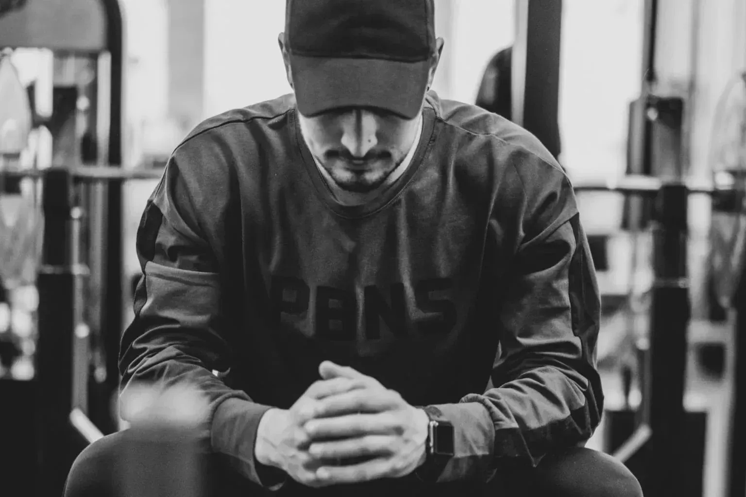 A person wearing a baseball cap and long-sleeved shirt sitting with hands clasped in a gym, captured in black and white.
