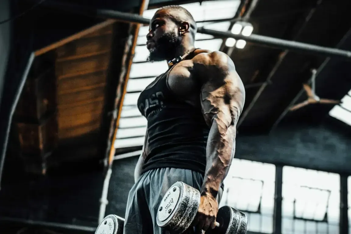 A person with a muscular build stands in a gym, holding dumbbells and looking off-camera.