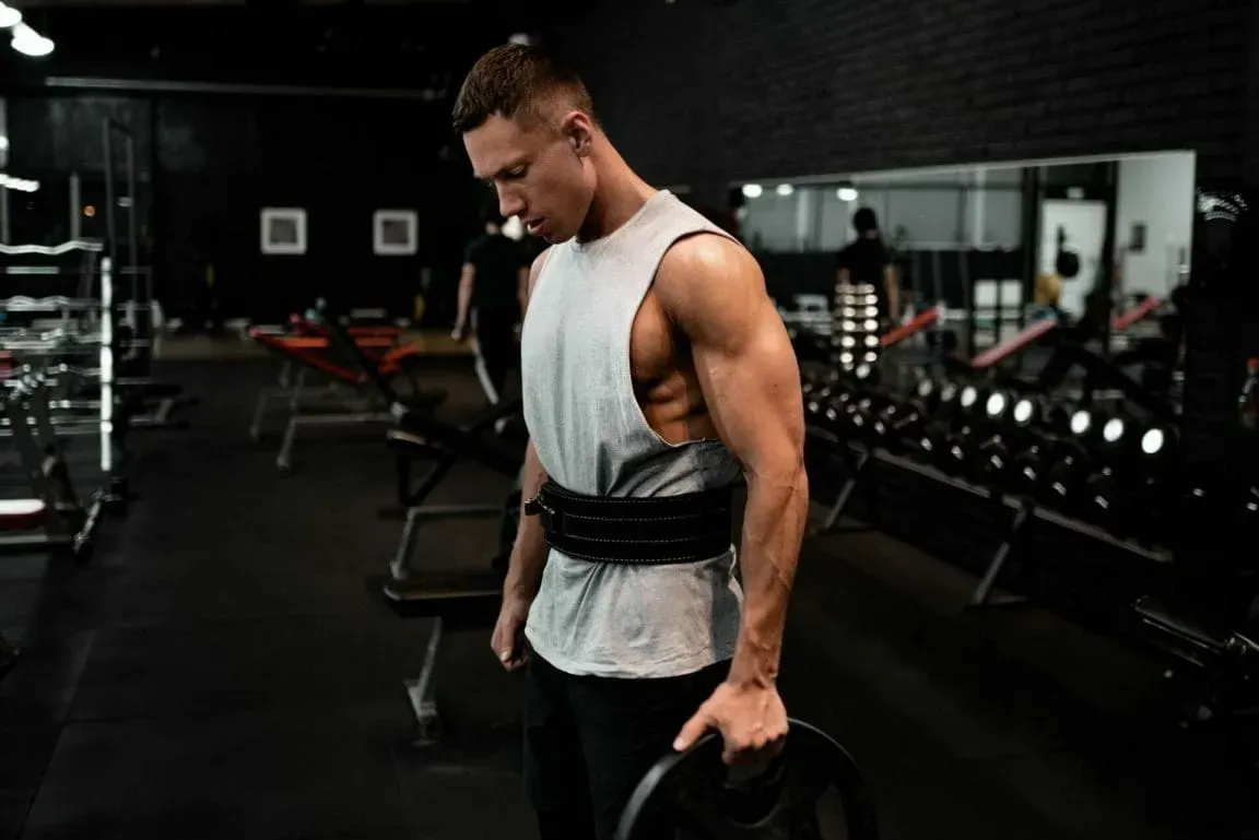 A focused individual in a gym wearing a lifting belt and holding a weight plate in a dimly lit, industrial setting.
