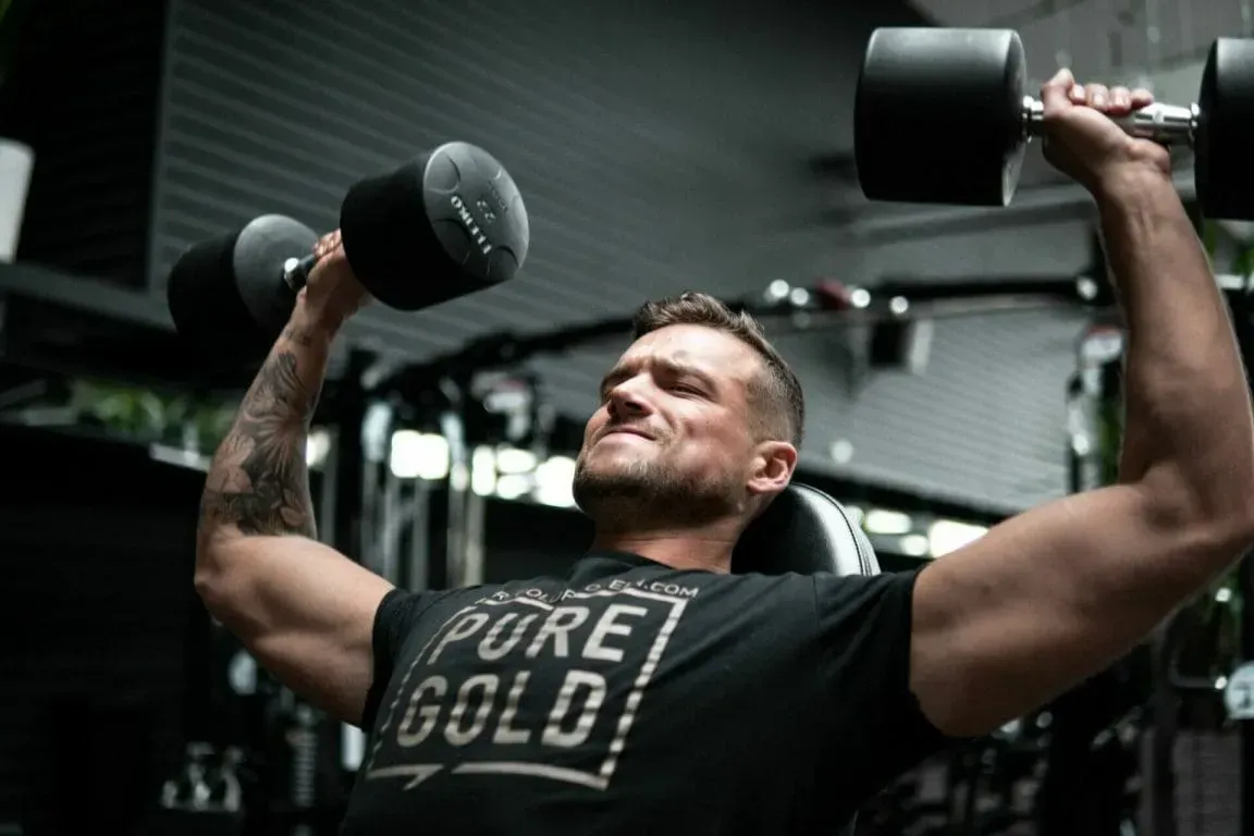 A person with a tattoo on their arm performing a seated dumbbell overhead press with a focused expression in a gym.