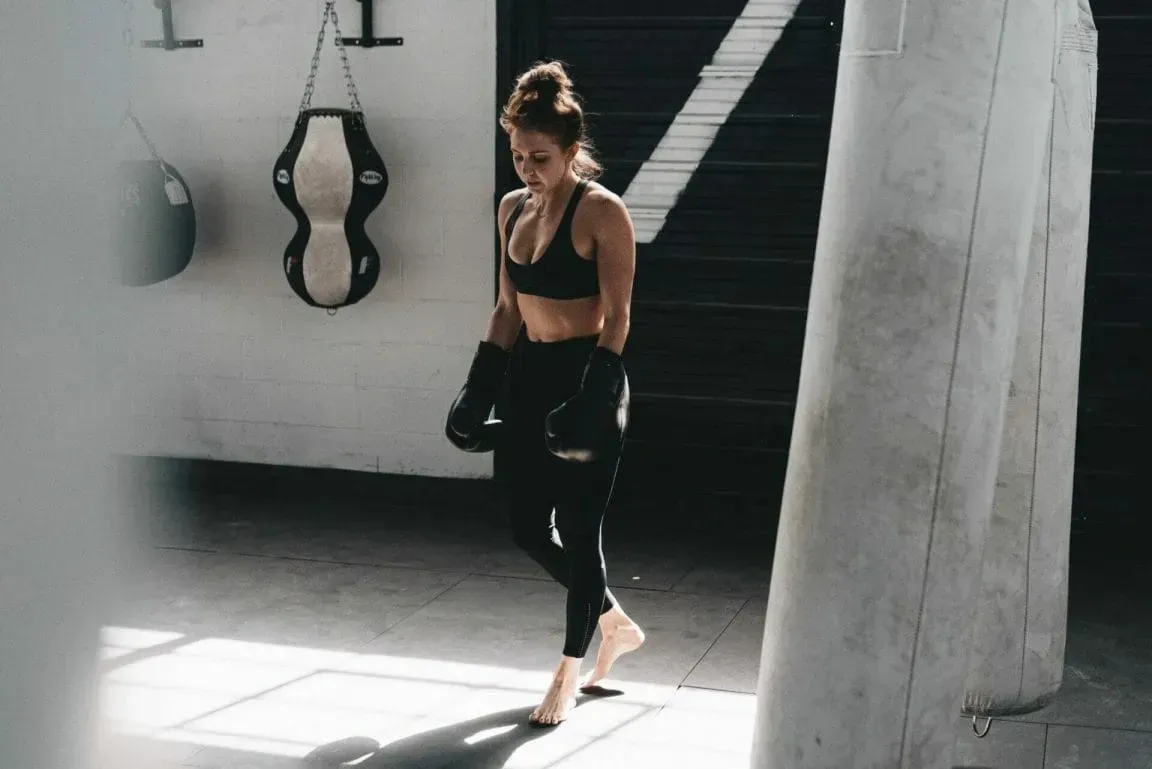 A person wearing a sports bra and leggings with boxing gloves stands in a gym near hanging punching bags.