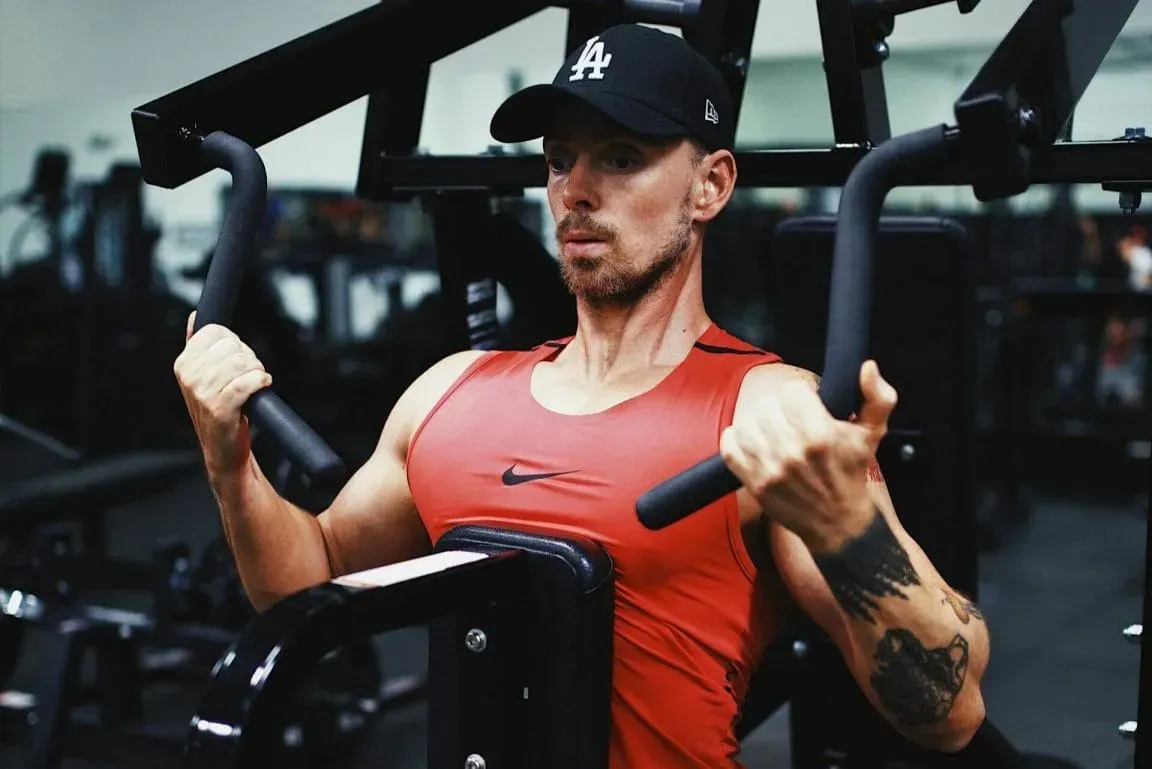 A person in a red Nike tank top and black baseball cap exercising on a chest press machine in a gym.