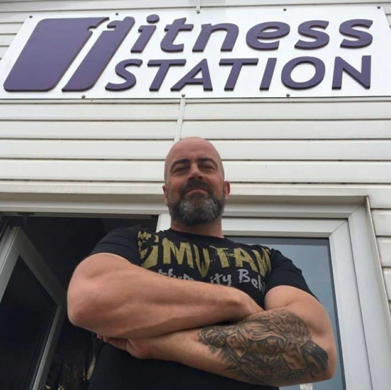 A person with a beard and tattoos stands with arms crossed in front of a building with a 