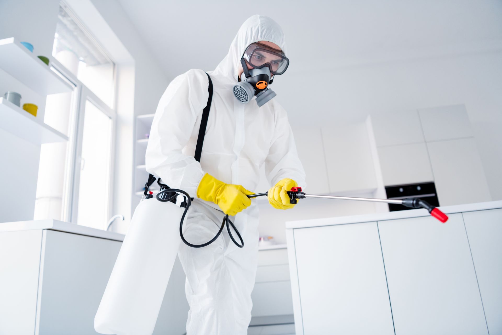 Person in white hazmat suit and respirator spraying disinfectant in a bright white room
