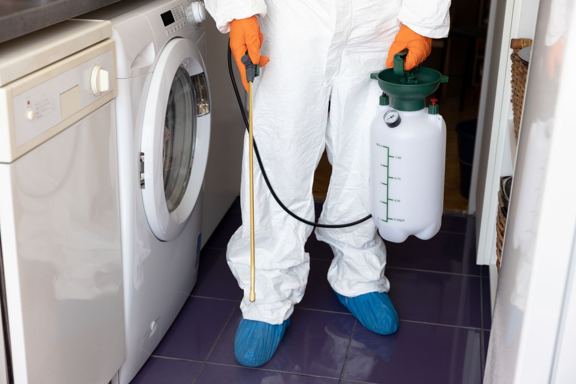 Technician in white coveralls using a sprayer beside a washing machine in a narrow laundry room.