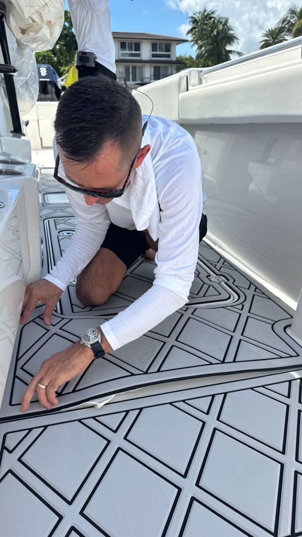 A person in a white long-sleeve shirt and black shorts kneels on a white boat deck with geometric black and white pattern