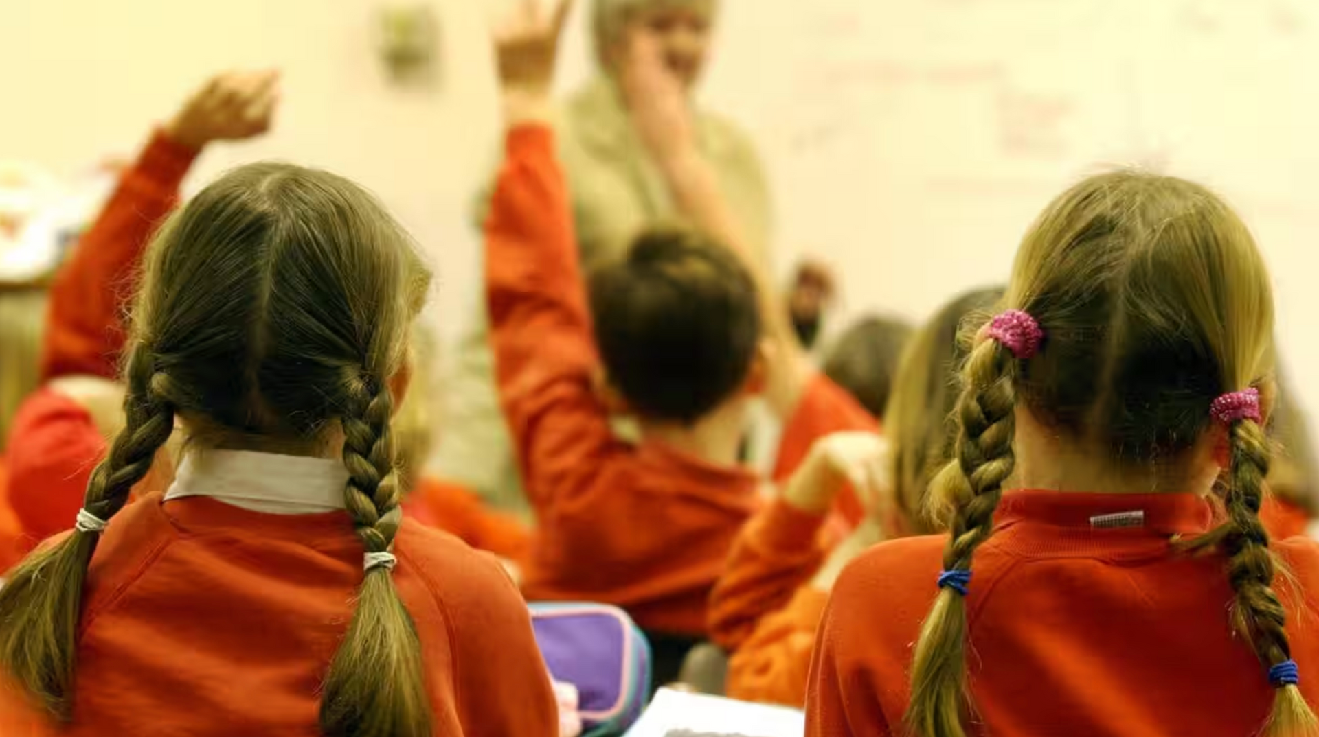 Children in red school uniforms with hands raised, classroom setting. Teacher in the background.