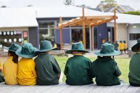 Children in green and yellow school uniforms sit on a wooden bench, facing away from the camera, at a school.