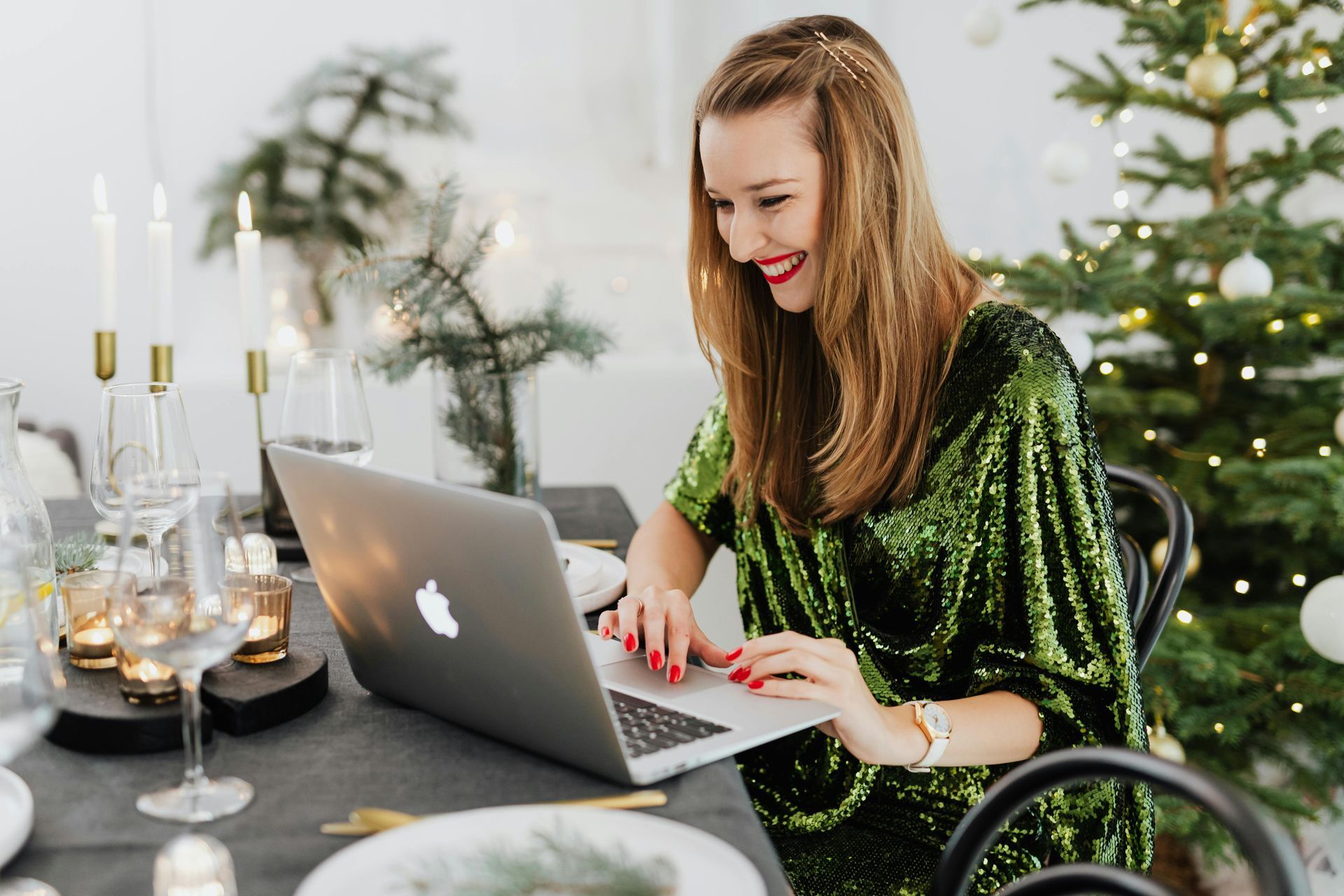 Woman in green sequin dress smiles at laptop at decorated Christmas table.
