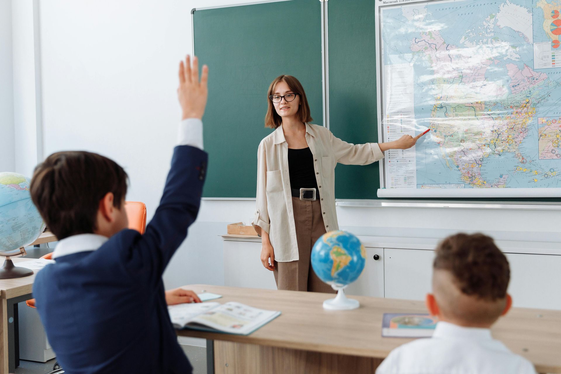 Teacher pointing at world map, student raising hand in classroom.
