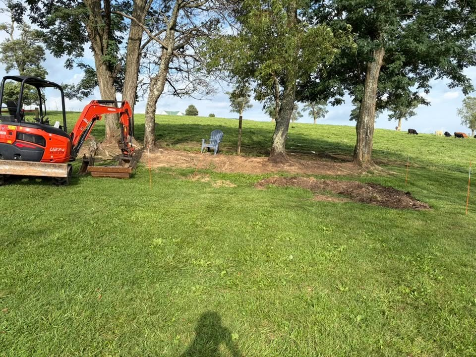 Orange excavator clearing grass near trees in a green field by a lake