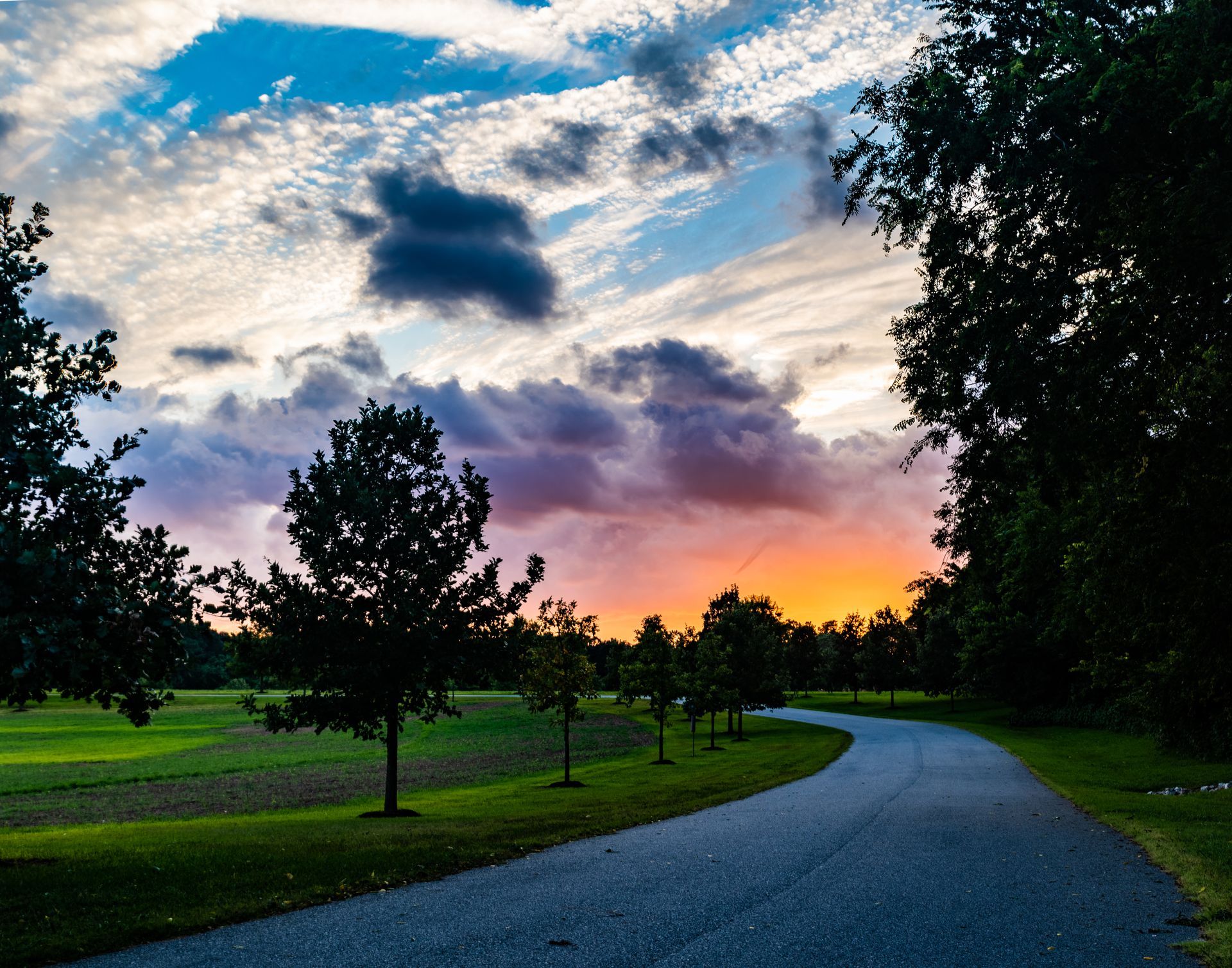 Gravel path curves through a park at sunset. Orange and purple hues illuminate the sky with trees lining the sides.
