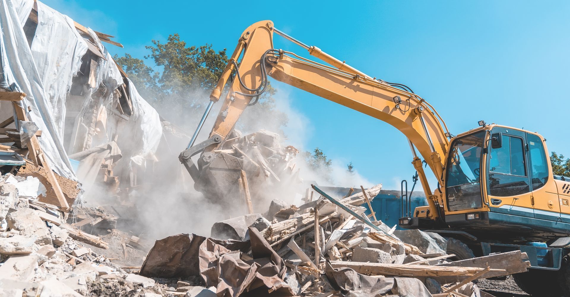 Yellow excavator demolishing a building, kicking up dust against a blue sky.