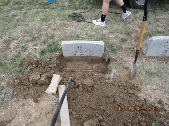 Fresh grave being dug with a shovel in a cemetery, with a person’s legs visible nearby.