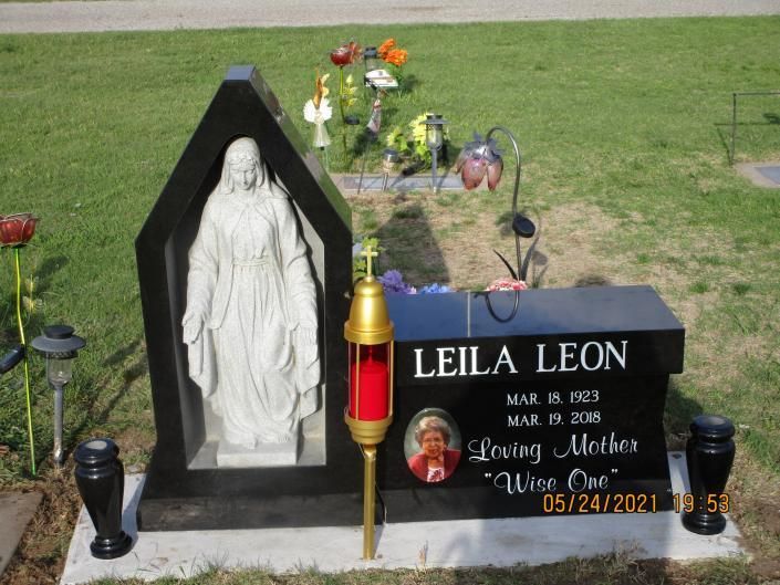Gravestone memorial with white statue, flowers, and black plaque reading “LEILA LEON” on grass.