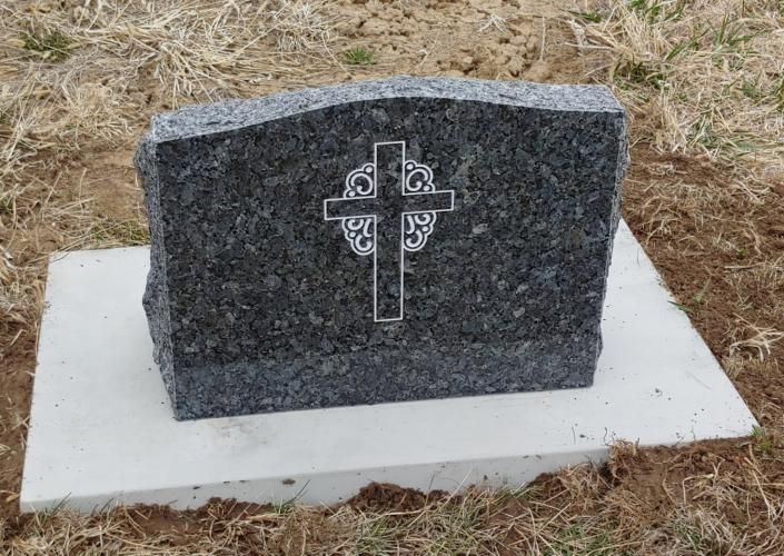 Dark granite memorial marker with a cross on a white concrete base in dry grass