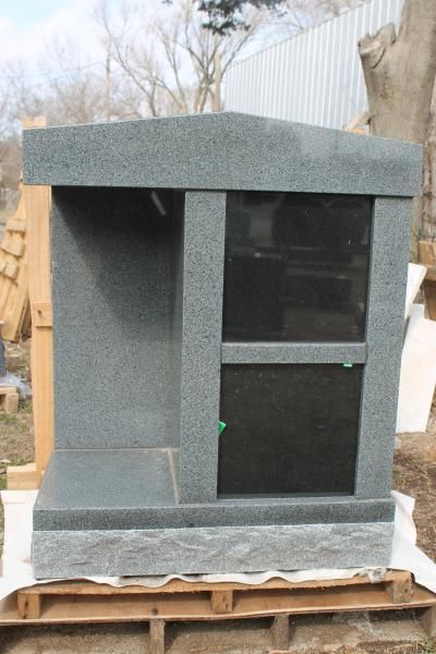 Gray stone memorial tombstone with black glass front, sitting on a wooden pallet outdoors.