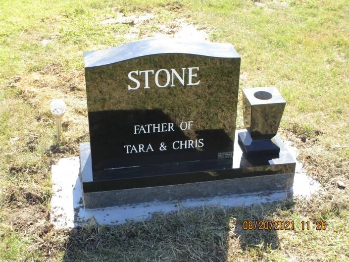Black gravestone reading “STONE, FATHER OF TARA & CHRIS” on a grassy cemetery plot.