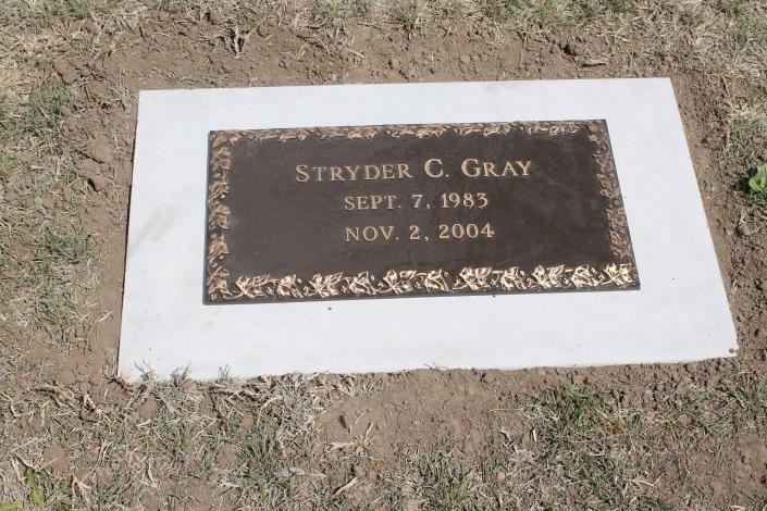 Gravestone plaque on a white concrete marker in dry grass, reading “Stryder C. Gray.”