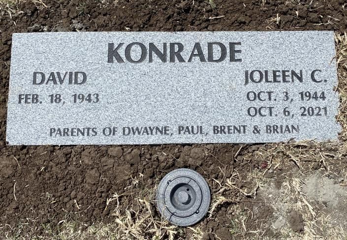 Gravestone for Konrade and Joleen C. David in dirt, with engraved names and dates.