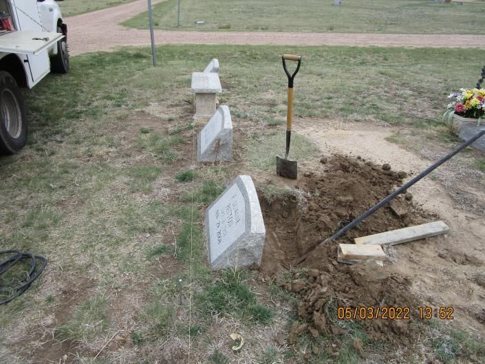 Fresh grave site with white markers, open dirt plot, and a shovel in a grassy cemetery area