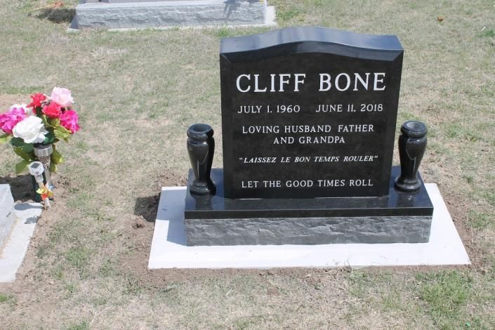 Black granite gravestone reading “CLIFF BONE” with flowers beside it in a cemetery.