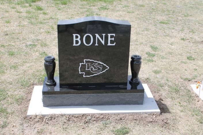 Black granite gravestone with “BONE” and a football logo on a white base in a grassy cemetery area