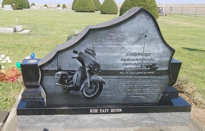 Black motorcycle memorial tombstone in a cemetery, with engraved text and grassy background