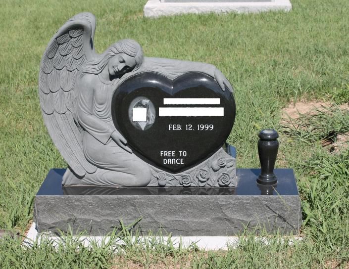 Black heart-shaped gravestone with angel carving and “FEB 12, 1999” in a grassy cemetery