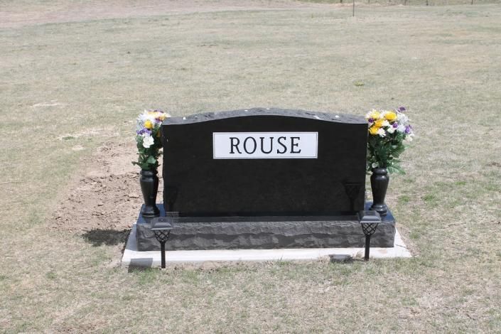 Black memorial headstone labeled “ROUSE” with flower arrangements on a grassy lawn