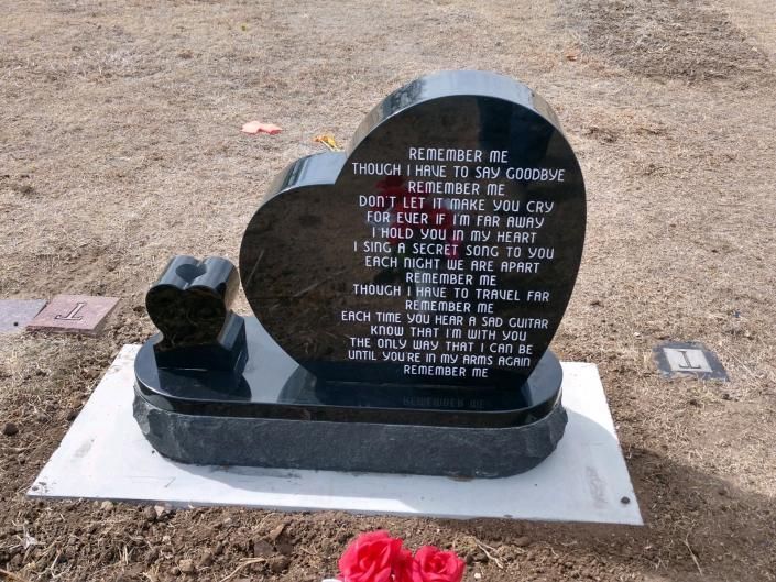 Black memorial headstone with engraved text and red flowers on a concrete base in a dry cemetery.