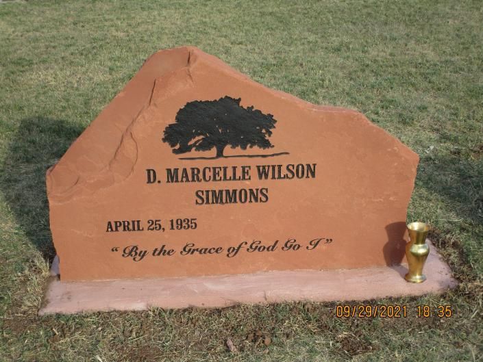 Brown gravestone in grass with engraved name and date, topped by a black silhouette and small gold vase
