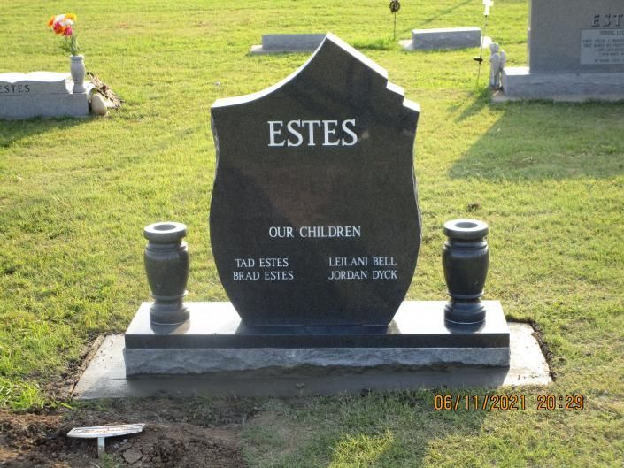 Black gravestone in a grassy cemetery marked “ESTES” with two small vases on either side
