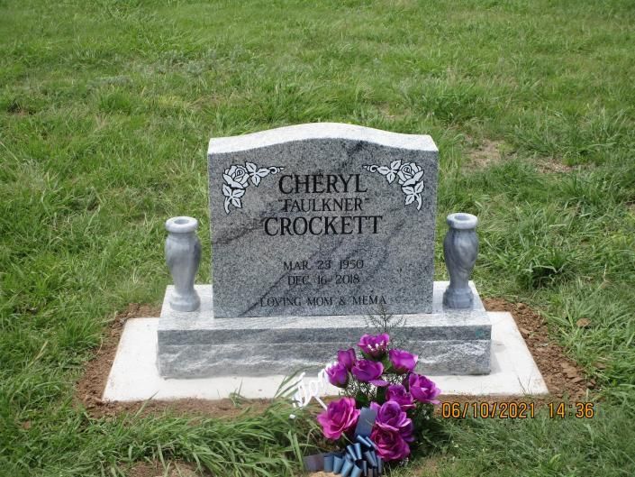 Gravestone in a grassy cemetery, with purple flowers placed at the base.