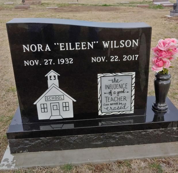 Black memorial gravestone for Nora “Eileen” Wilson with schoolhouse engraving and pink flowers on the right.