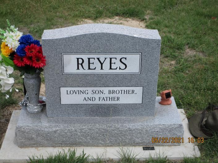 Gray gravestone marked “REYES” with flowers beside it on grass